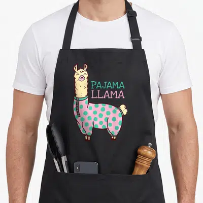 Pajama alpaca apron - Cartoon llama in pink teal polka dot pajamas next to stacked text — shown on a black cotton canvas apron against a white background