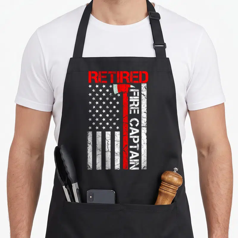 Captain firefighter cooking apron - Red and white distressed flag with axe and RETIRED FIRE CAPTAIN text — shown on a black cotton canvas apron against a white background