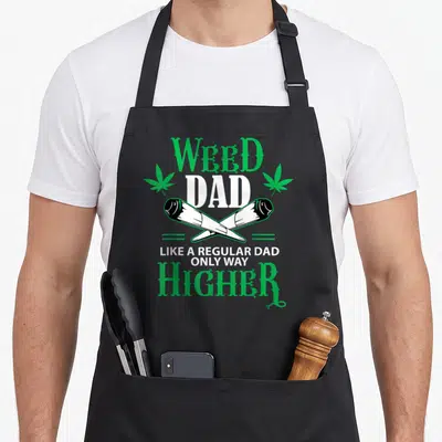 Weed pumpkin apron - Stacked Weeds Dad text with two cannabis leaves and crossed joints in green and white — shown on a black cotton canvas apron against a white background