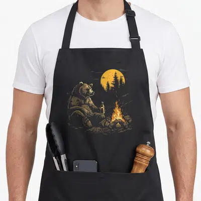 Drinking bear apron - Realistic gold bear holding bottle sitting near campfire with sun and trees — shown on a black cotton canvas apron against a white background