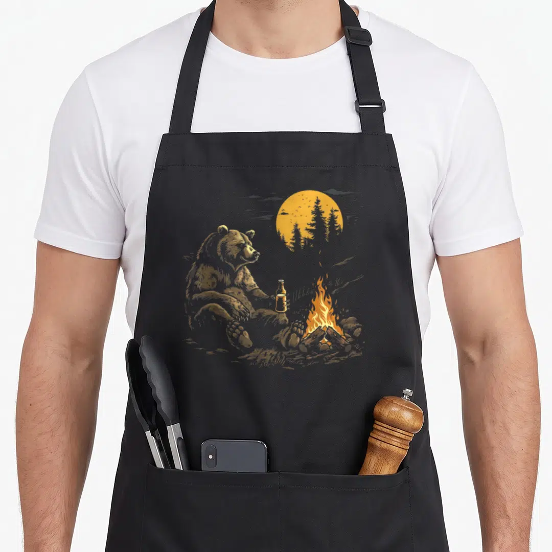 Drinking bear apron - Realistic gold bear holding bottle sitting near campfire with sun and trees — shown on a black cotton canvas apron against a white background