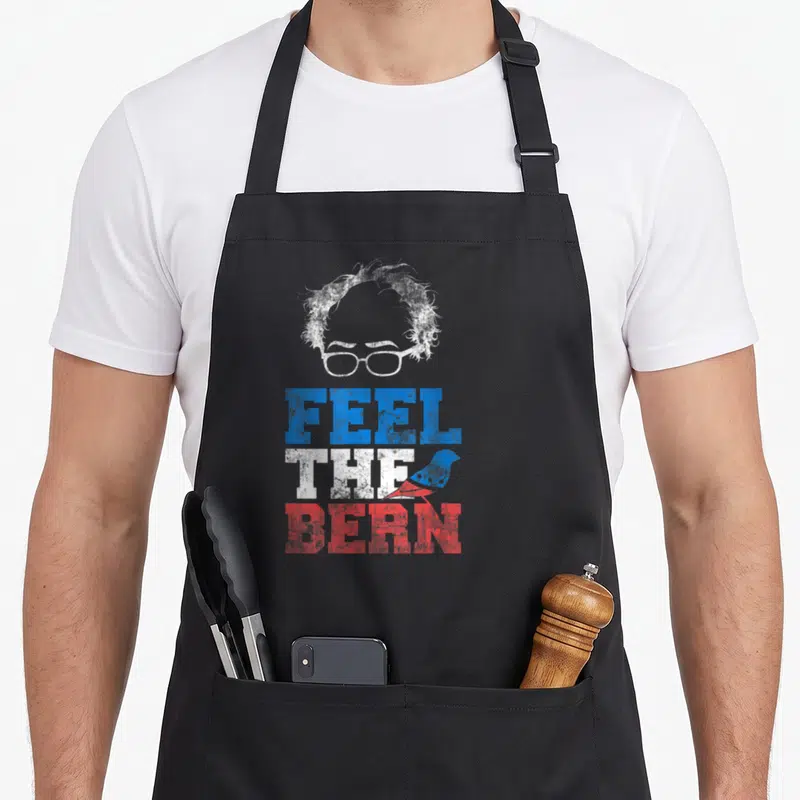 Bernie bird apron - Stylized head glasses FEEL THE BERN text blue white red distressed sparrow — shown on a black cotton canvas apron against a white background