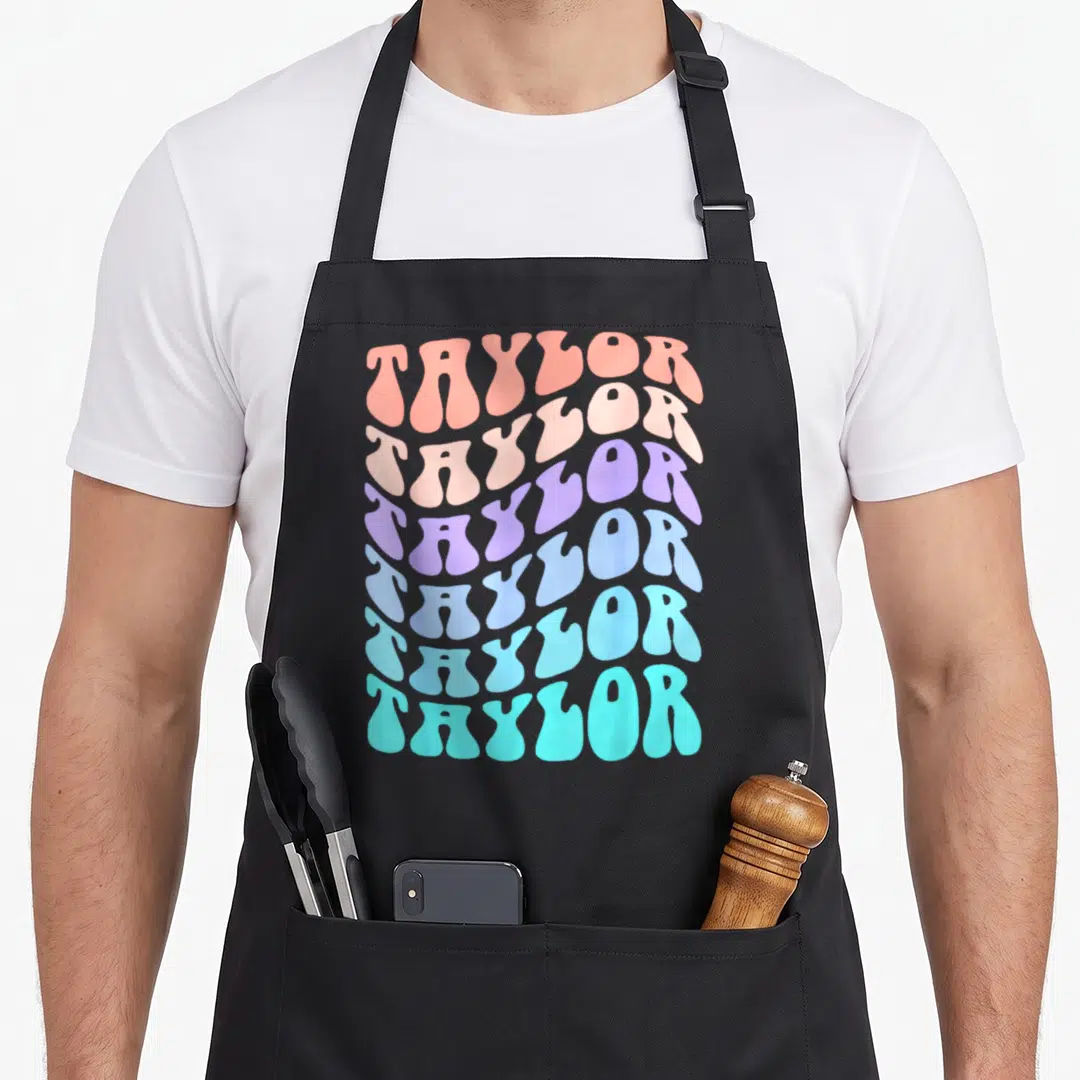 Taylor groovy birthday apron - Six stacked wavy lines of text reading TAYLOR in coral, pink, purple, blue, and turquoise — shown on a black cotton canvas apron against a white background
