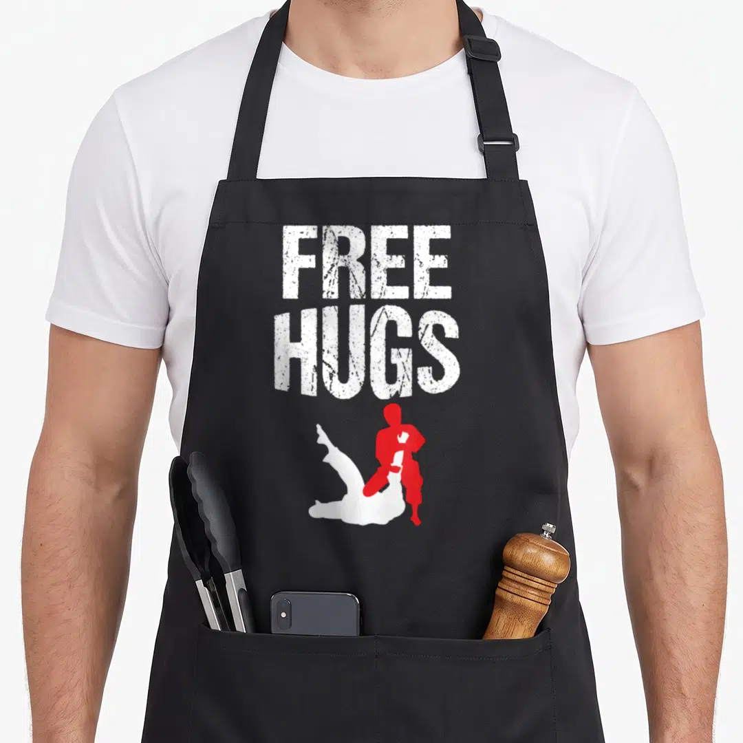 Free jitsu apron - Distressed white FREE HUGS text above red and white wrestler silhouettes — shown on a black cotton canvas apron against a white background
