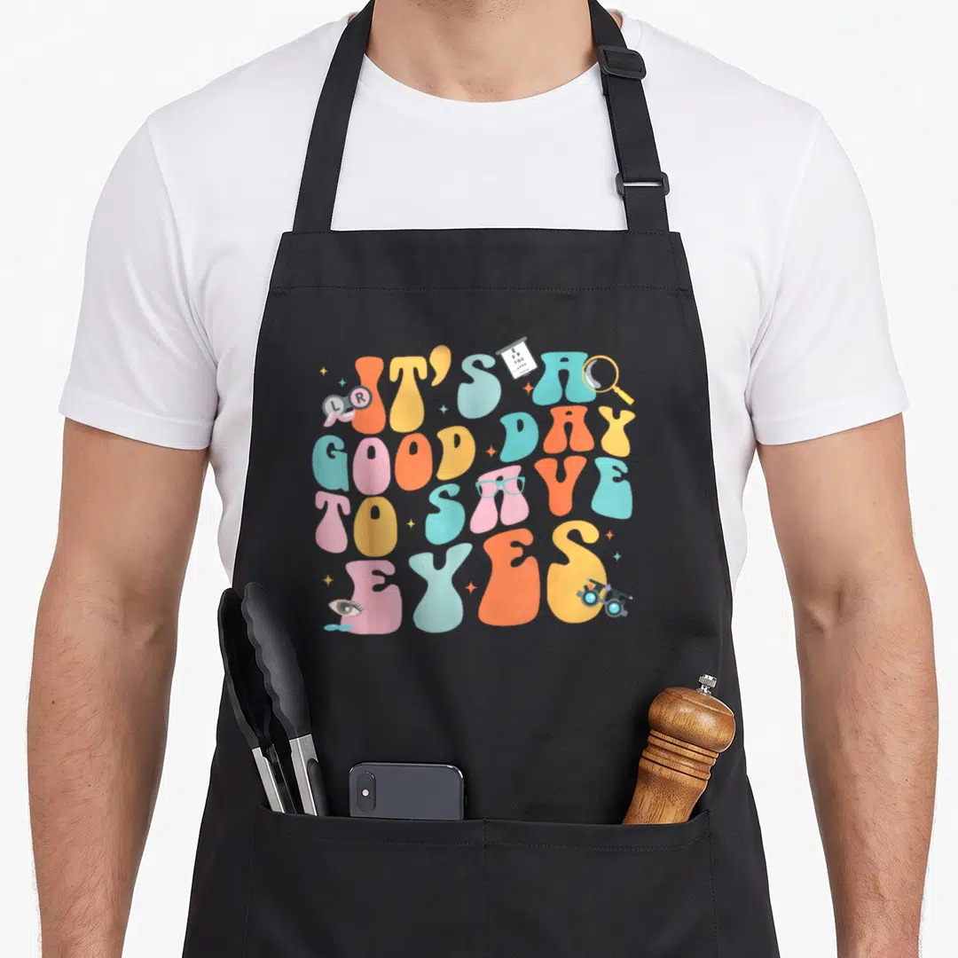 Save surgeon apron - Retro wavy text IT'S A GOOD DAY TO SAVE EYES with eye chart, glasses, and phoropter in orange — shown on a black cotton canvas apron against a white background