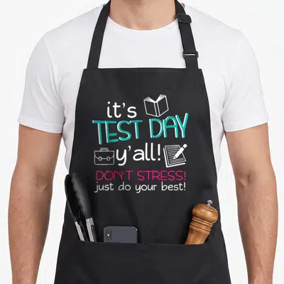 Test testing teacher apron - Vertical typography reading It's Test Day with book briefcase notebook and pencil icons — shown on a black cotton canvas apron against a white background