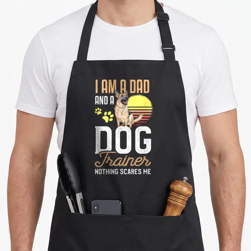Father dog trainer apron - German Shepherd sitting before retro sun with stacked text I AM A DAD AND A DOG Trainer — shown on a black cotton canvas apron against a white background