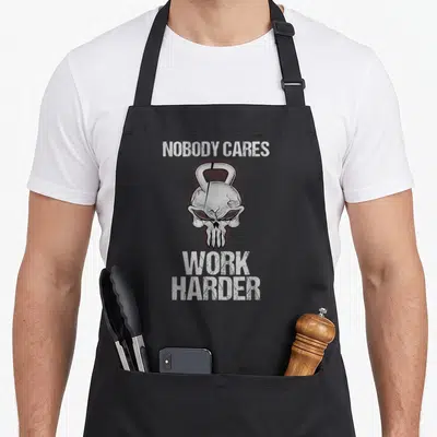 Nobody gym class apron - Distressed skull merged with kettlebell and white text on black background — shown on a black cotton canvas apron against a white background