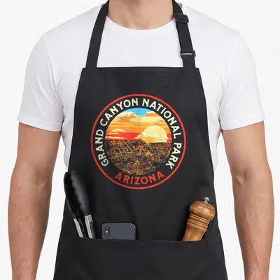 Grand canyon 80s apron - Circular red border badge with GRAND CANYON NATIONAL PARK text and Arizona sunset canyon — shown on a black cotton canvas apron against a white background