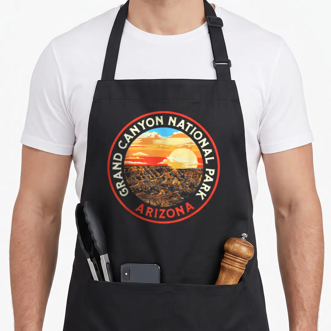 Grand canyon 80s apron - Circular red border badge with GRAND CANYON NATIONAL PARK text and Arizona sunset canyon — shown on a black cotton canvas apron against a white background