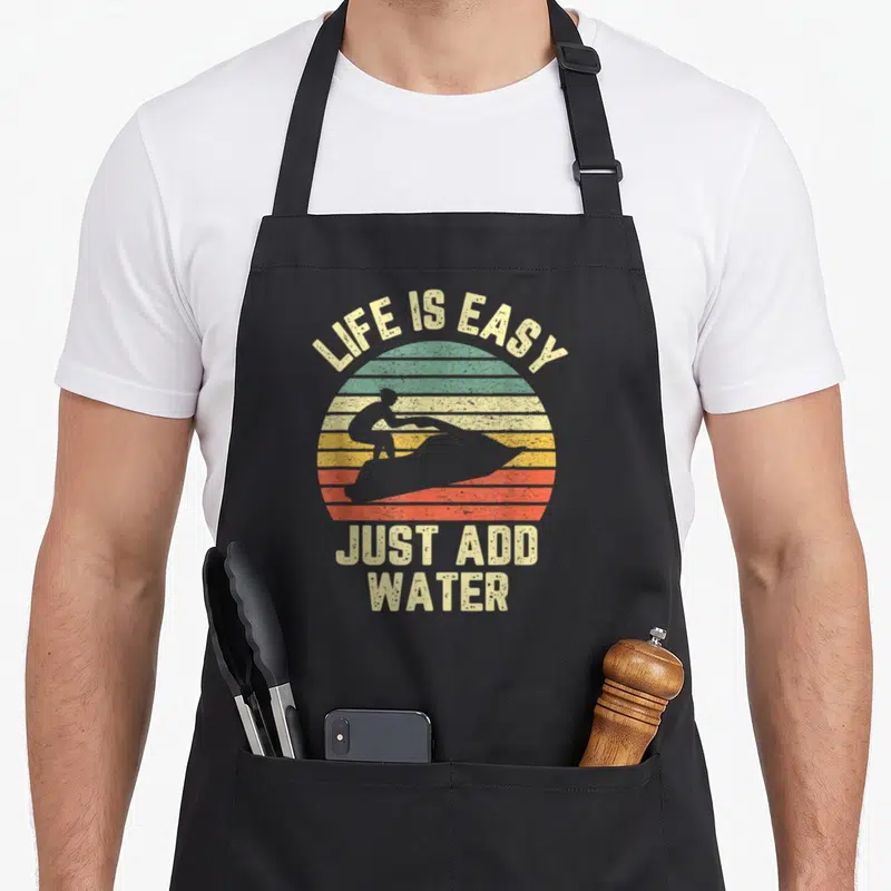 Jet easy apron - Retro striped sun with jet ski silhouette and LIFE IS EASY JUST ADD WATER text — shown on a black cotton canvas apron against a white background