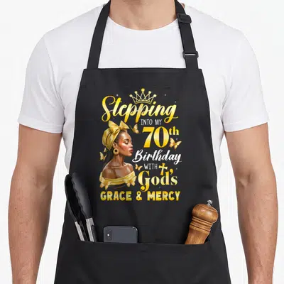 Afro 70th birthday apron - Woman profile with crown and butterflies next to Stepping Into My 70th Birthday With God's — shown on a black cotton canvas apron against a white background