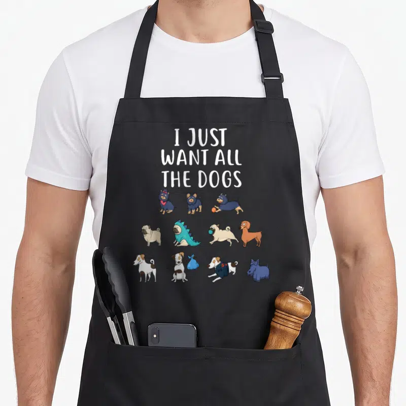 General dog apron - Cartoon dogs in three rows below stacked text I JUST WANT ALL THE DOGS in white blue tan brown — shown on a black cotton canvas apron against a white background