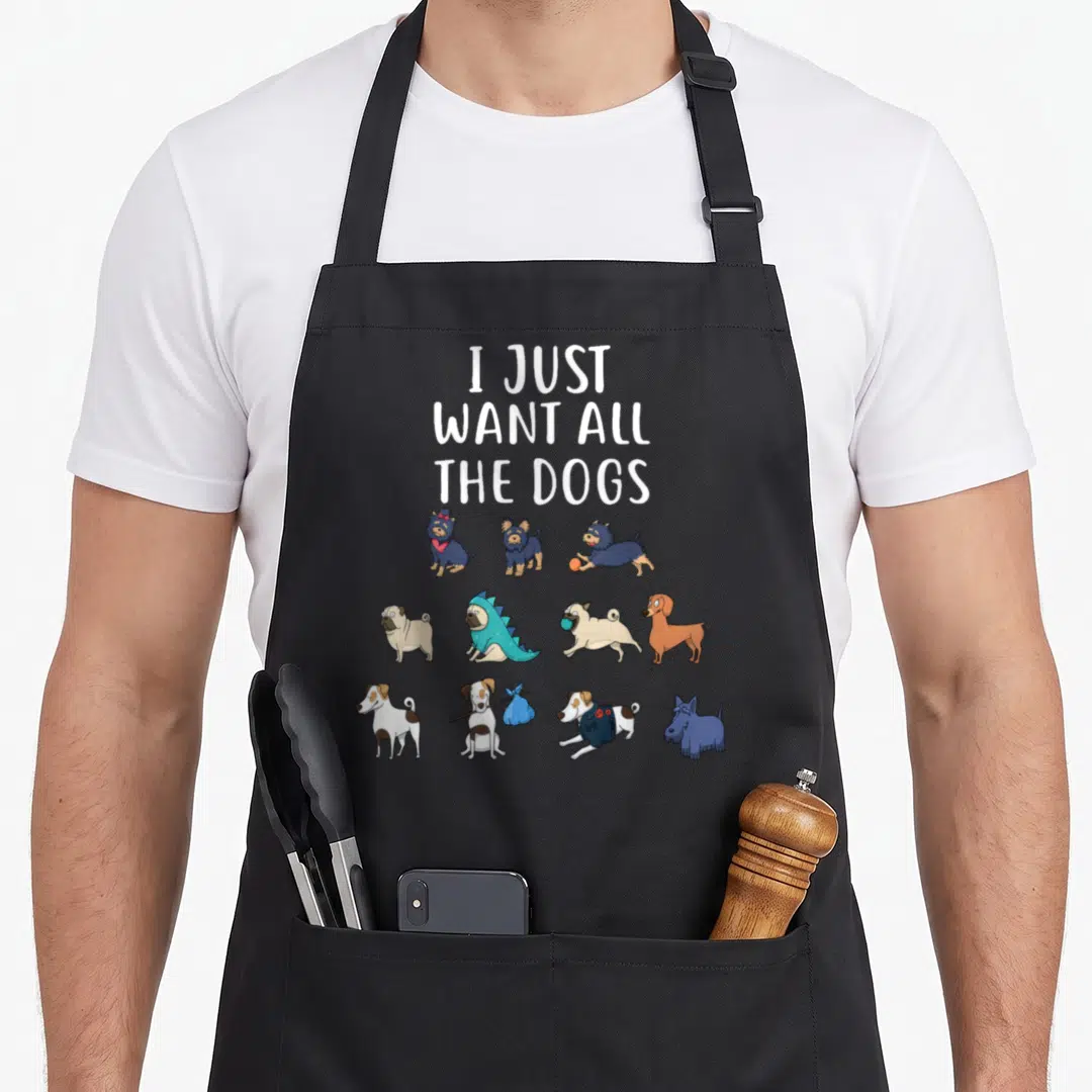 General dog apron - Cartoon dogs in three rows below stacked text I JUST WANT ALL THE DOGS in white blue tan brown — shown on a black cotton canvas apron against a white background