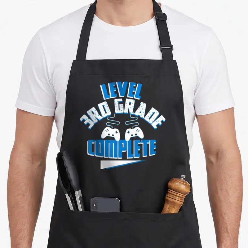 School game day apron - LEVEL 3RD GRADE COMPLETE text with two blue white game controllers — shown on a black cotton canvas apron against a white background