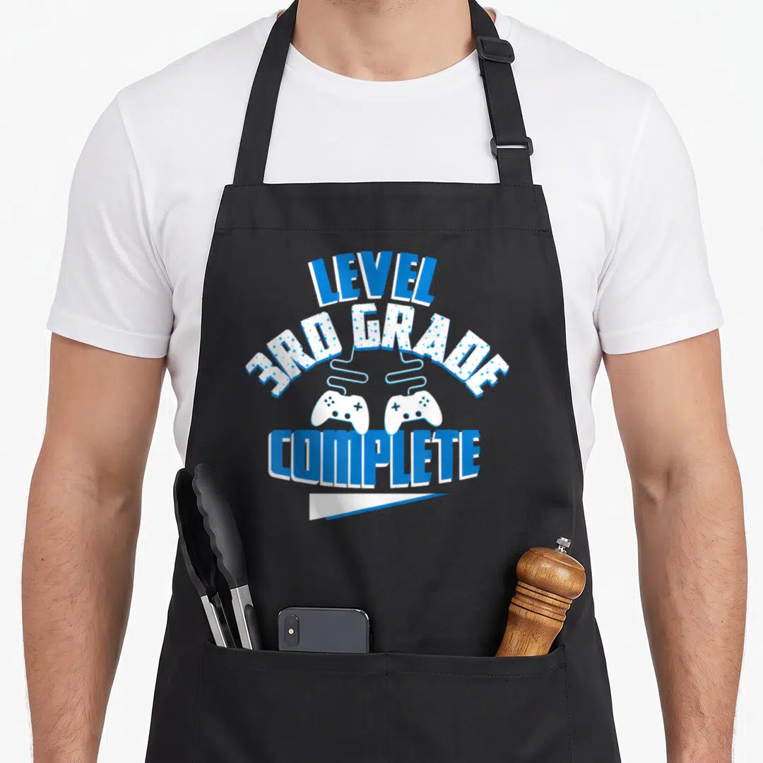 School game day apron - LEVEL 3RD GRADE COMPLETE text with two blue white game controllers — shown on a black cotton canvas apron against a white background