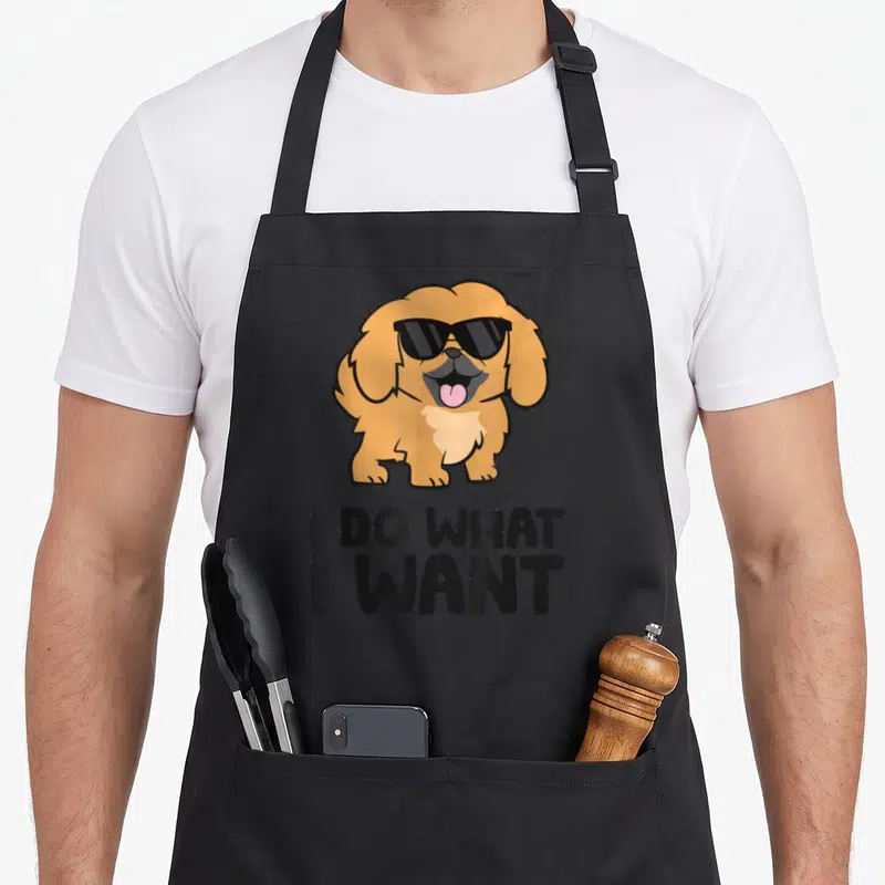 Funny pekingese dog apron - Tan cartoon dog in black sunglasses with pink tongue above stacked black text reading I DO — shown on a black cotton canvas apron against a white background
