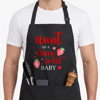 Aunt strawberry apron - Watercolor strawberries and hearts surround stacked red and white text reading aunt of a berry — shown on a black cotton canvas apron against a white background