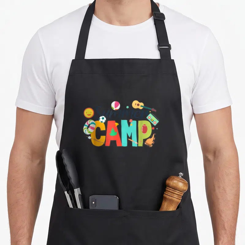Summer director camp chef apron - Cartoon Summer CAMP text with boom box, guitar, balls, marshmallow stick — shown on a black cotton canvas apron against a white background