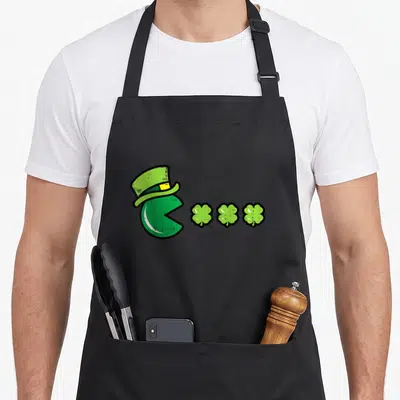 Eating st patrick day aprons - Green Pac-Man top hat facing three green shamrocks pixel art — shown on a black cotton canvas apron against a white background