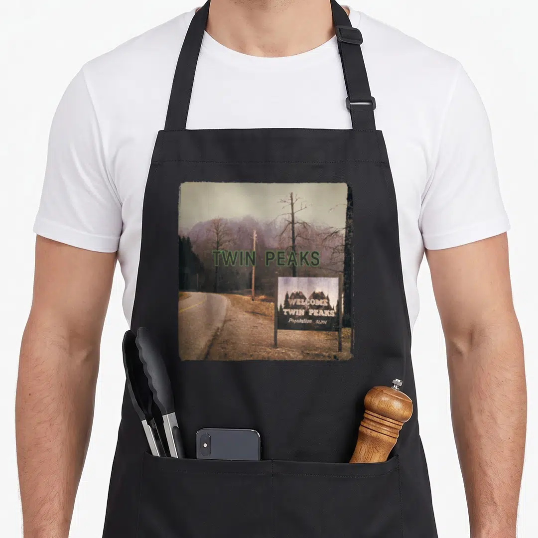 Limit twin peaks apron - Vintage road photo with Twin Peaks welcome sign and population 51,201 text — shown on a black cotton canvas apron against a white background