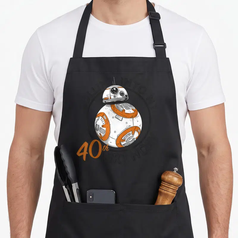 Rollin 40th birthday apron - BB-8 robot in grey circle with Rollin Into My 40th Birthday text — shown on a black cotton canvas apron against a white background