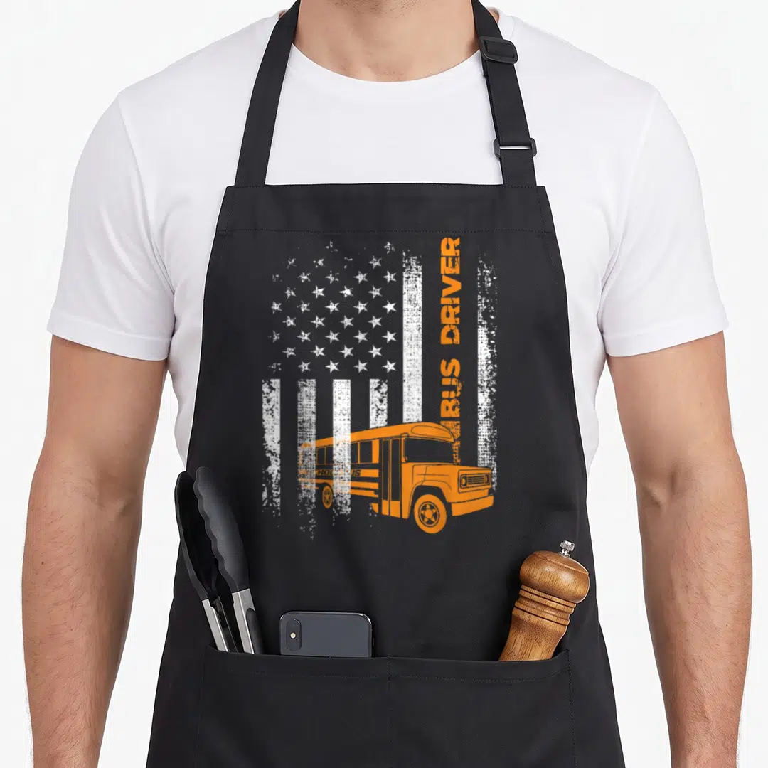 Bus american flag apron - Distressed flag background with orange school bus and vertical BUS DRIVER text — shown on a black cotton canvas apron against a white background