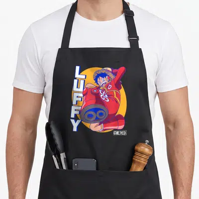 Egghead anime apron - Luffy in red suit with cannon arm and vertical text on orange circle — shown on a black cotton canvas apron against a white background