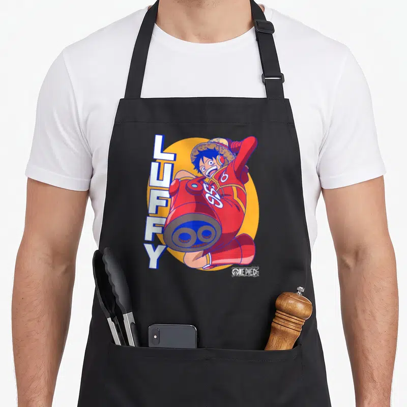 Egghead anime apron - Luffy in red suit with cannon arm and vertical text on orange circle — shown on a black cotton canvas apron against a white background