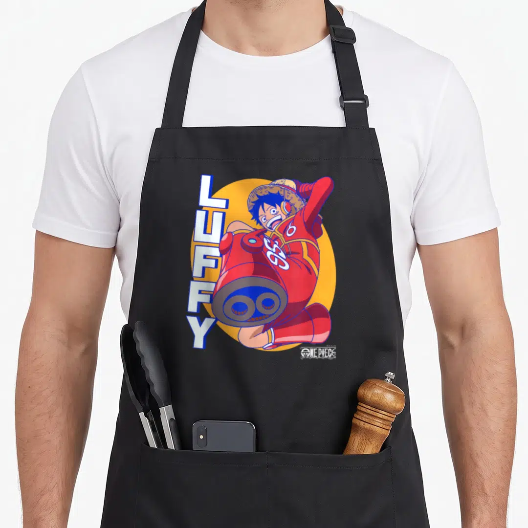 Egghead anime apron - Luffy in red suit with cannon arm and vertical text on orange circle — shown on a black cotton canvas apron against a white background