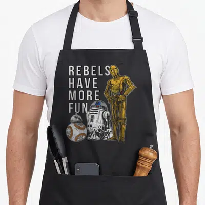 Droid droewors apron - C-3PO R2-D2 BB-8 robots with stacked text REBELS HAVE MORE FUN in gold and white — shown on a black cotton canvas apron against a white background