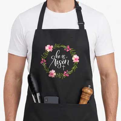Risen easter apron - he is Risen white script in pink flower wreath with cross below — shown on a black cotton canvas apron against a white background