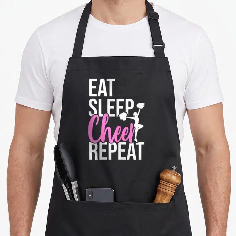 Eat gymnastics coach apron - Stacked EAT SLEEP Cheer REPEAT text with white cheerleader silhouette in pink and white — shown on a black cotton canvas apron against a white background