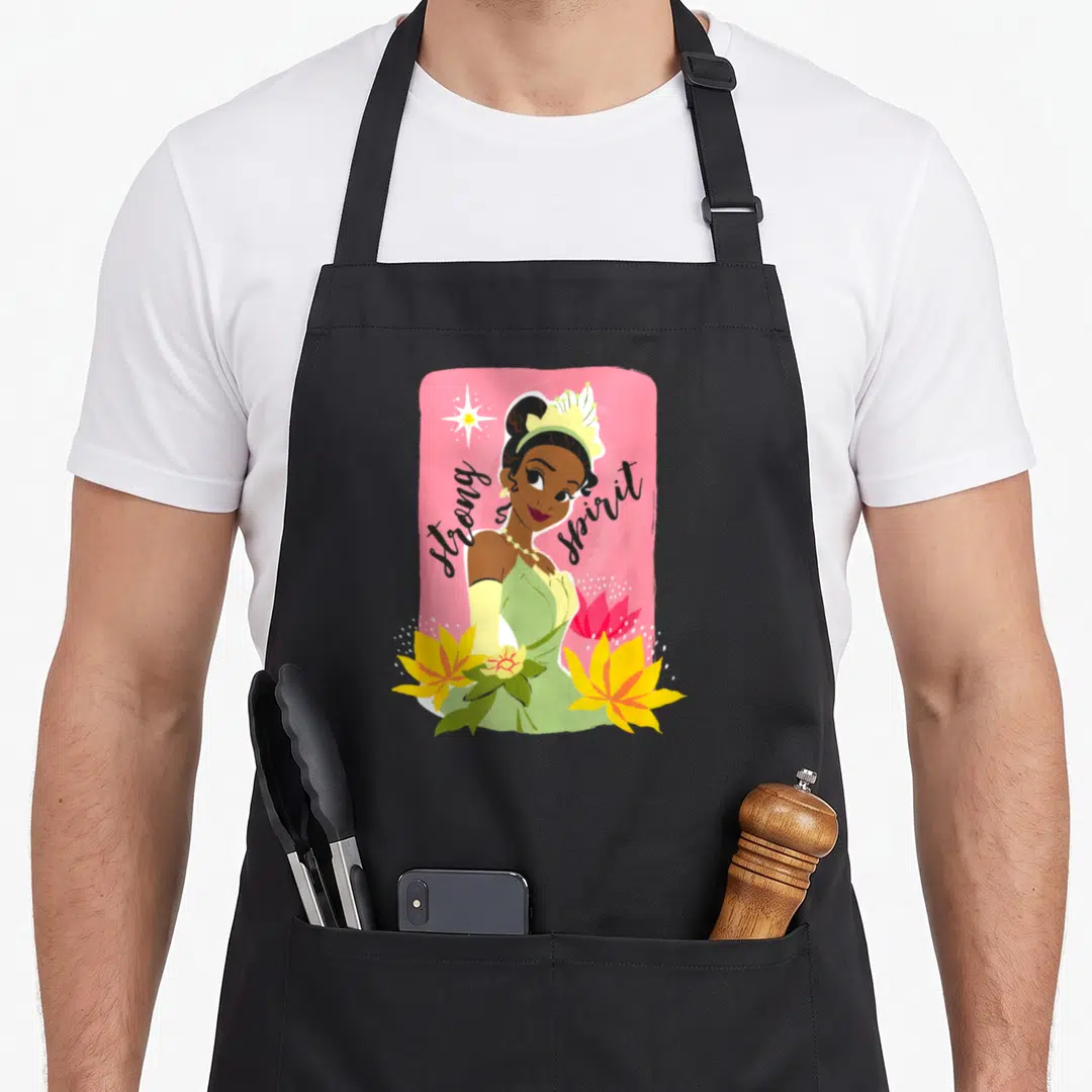 Free princess apron - Cartoon woman on pink background with strong spirit text, yellow flowers, and star — shown on a black cotton canvas apron against a white background