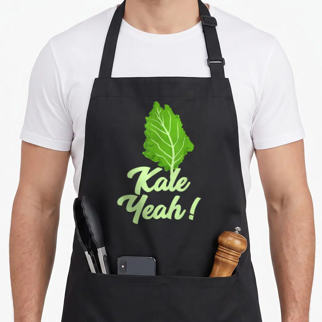 Pun vegan apron - Green cartoon kale leaf above light green script reading Kale Yeah! — shown on a black cotton canvas apron against a white background