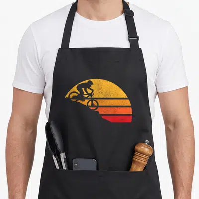 Mountain cycling apron - Grey cyclist silhouette against yellow orange and red striped sun — shown on a black cotton canvas apron against a white background