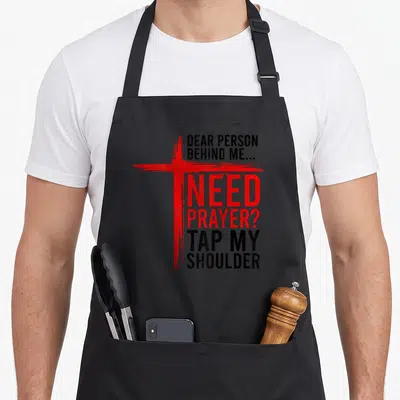 Dear shoulder apron - Distressed red cross next to stacked black and red text requesting prayer support — shown on a black cotton canvas apron against a white background