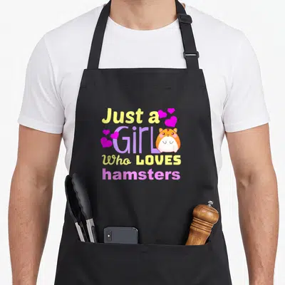 Funny loves hamsters apron - Cartoon hamster next to stacked yellow and purple text reading Just a Girl Who LOVES — shown on a black cotton canvas apron against a white background