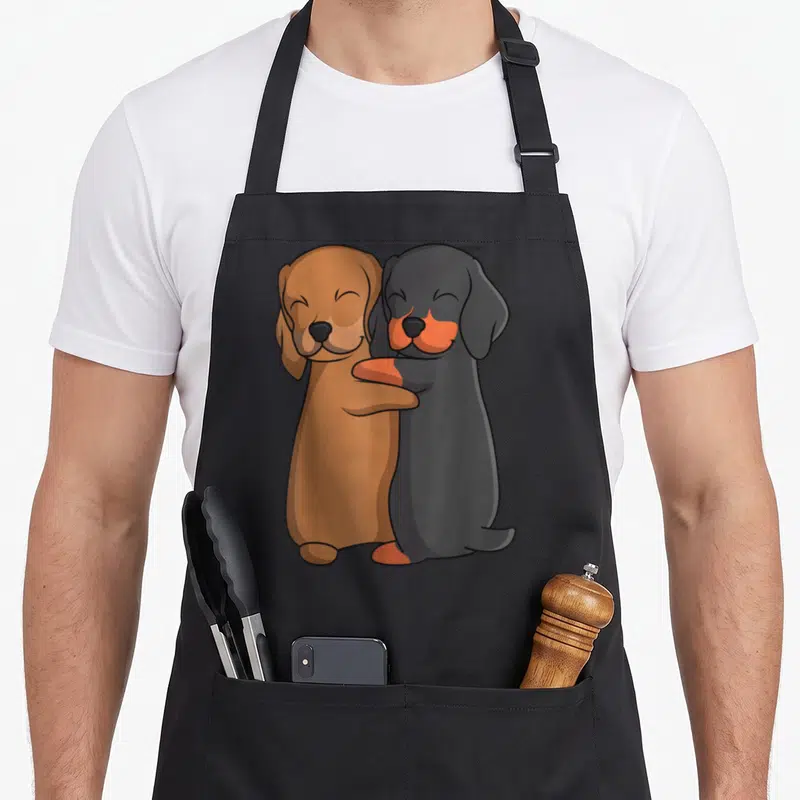 Weiner dog dachshund apron - Two cartoon dachshunds hugging, one brown and one black with orange accents — shown on a black cotton canvas apron against a white background