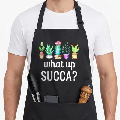 Succa cactus apron - Five watercolor succulents in pots above white text reading what up SUCCA — shown on a black cotton canvas apron against a white background