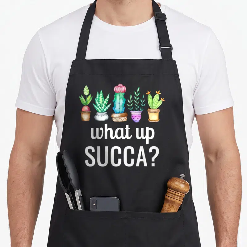 Succa cactus apron - Five watercolor succulents in pots above white text reading what up SUCCA — shown on a black cotton canvas apron against a white background
