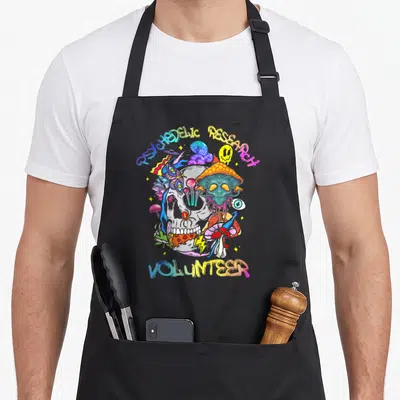 Research mushroom apron - Cartoon skull surrounded by mushrooms and alien with rainbow text reading psychedelic — shown on a black cotton canvas apron against a white background