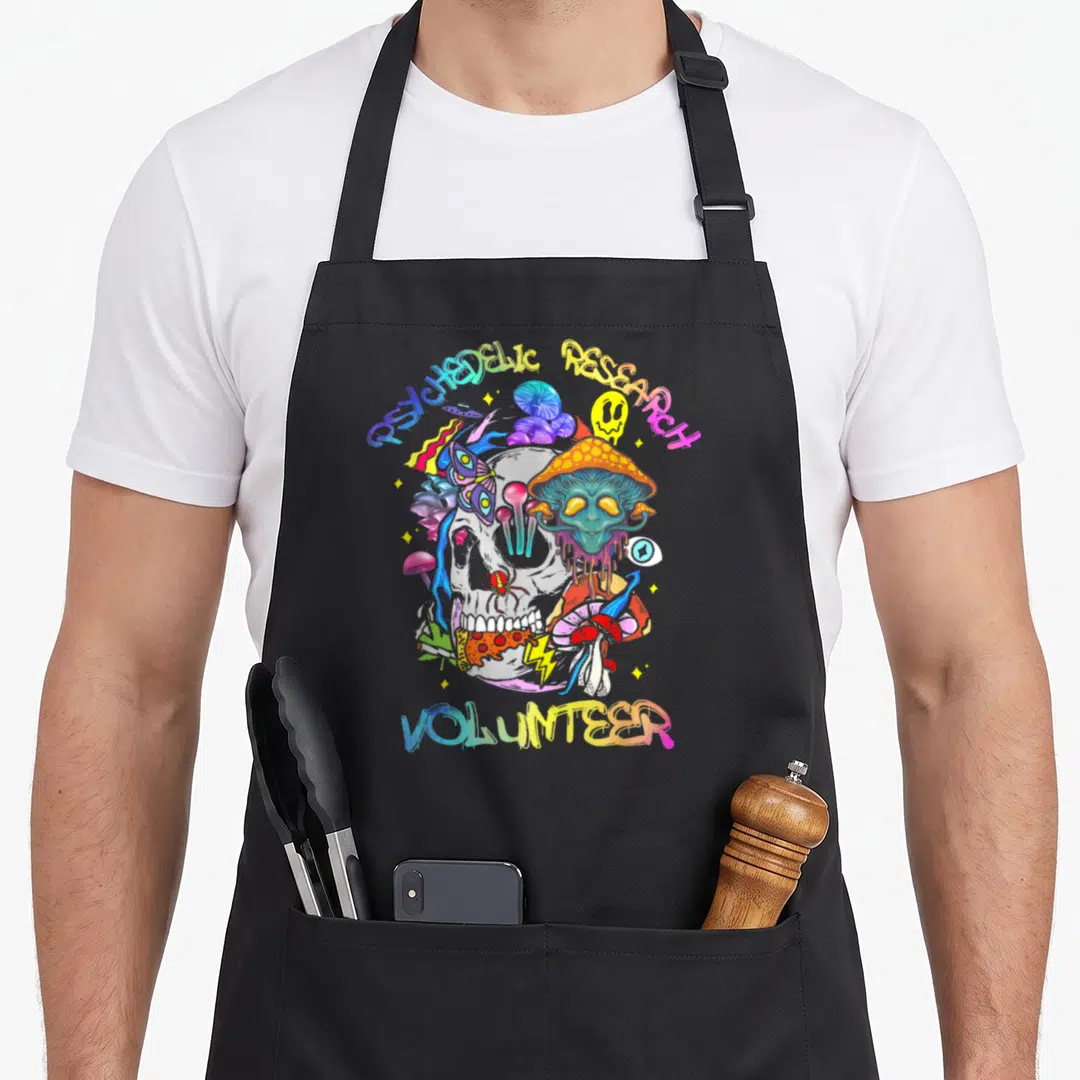 Research mushroom apron - Cartoon skull surrounded by mushrooms and alien with rainbow text reading psychedelic — shown on a black cotton canvas apron against a white background