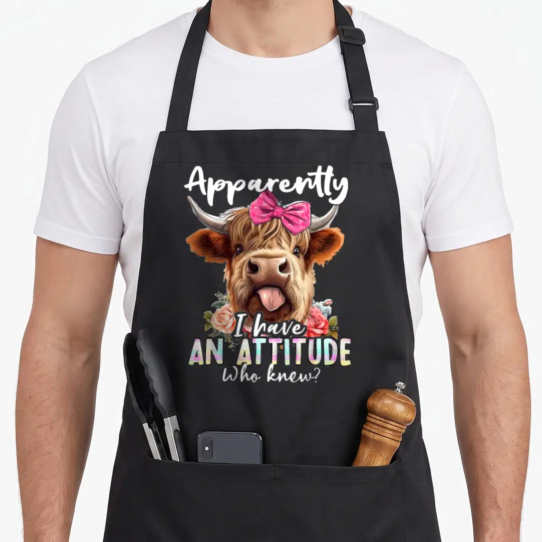 Apparently highland cow apron - Cartoon highland cow with pink bow and roses under text 'I have AN ATTITUDE Who knew' — shown on a black cotton canvas apron against a white background
