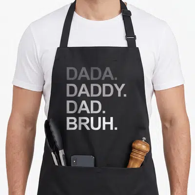 Bruh father day apron - Stacked grey text DADA DADDY DAD BRUH on black background — shown on a black cotton canvas apron against a white background