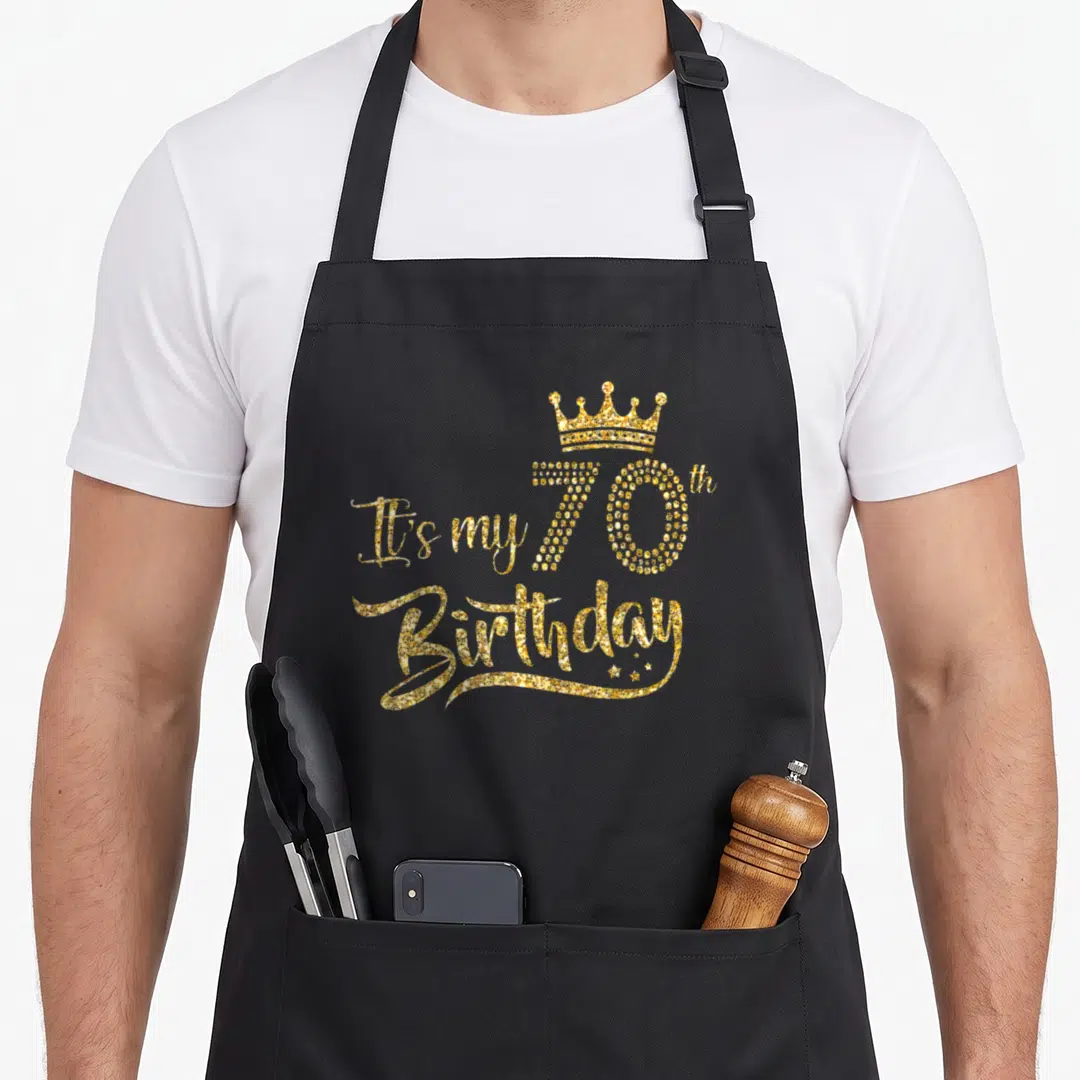 70th queen of baking apron - Gold glitter text It's my 70th Birthday with crown and three stars — shown on a black cotton canvas apron against a white background