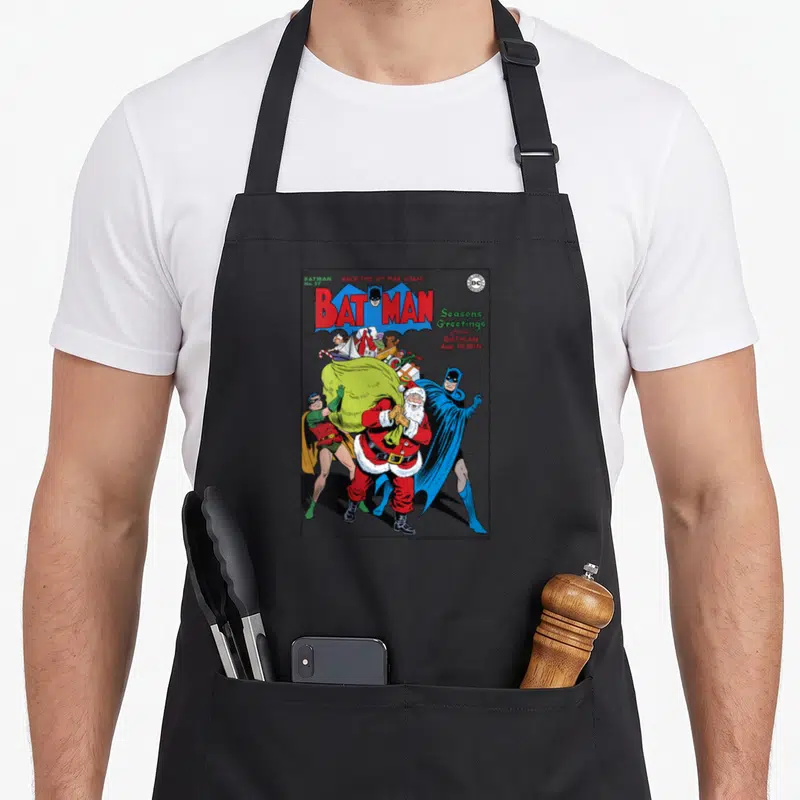 Season greeting christmas robin apron - Vintage Batman comic cover art featuring Santa Claus and a green toy sack — shown on a black cotton canvas apron against a white background