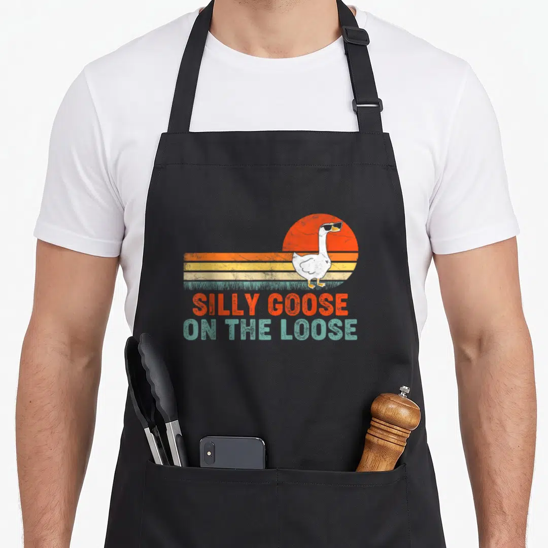 Funny silly goose apron - White goose in sunglasses on orange yellow teal retro sunset stripes with stacked text — shown on a black cotton canvas apron against a white background
