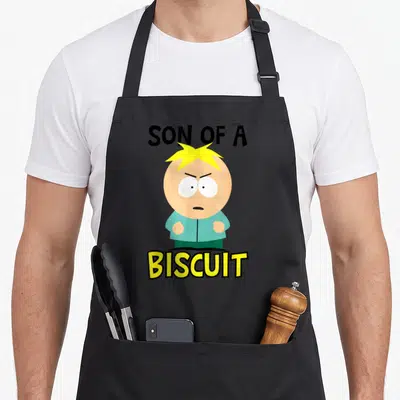 Son butter apron - Cartoon boy with yellow hair between SON OF A and BISCUIT text — shown on a black cotton canvas apron against a white background