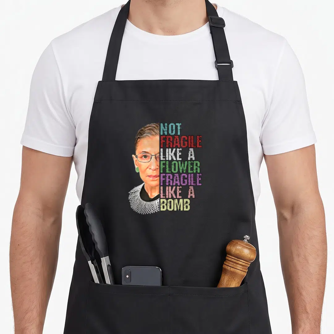 Fragile novelty apron - Woman portrait with stacked text Not Fragile Like a Flower Fragile Like a Bomb in multi-color — shown on a black cotton canvas apron against a white background